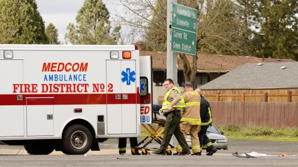 White ambulance and three first responders in yellow vests and firefighting suit wheel gurney into ambulance at car crash site at small intersection.
