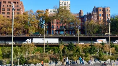View of joggers on Brooklyn Bridge Park trail in foreground with Brooklyn-Queens Expressway traffic in background and brick buildings behind the highway
