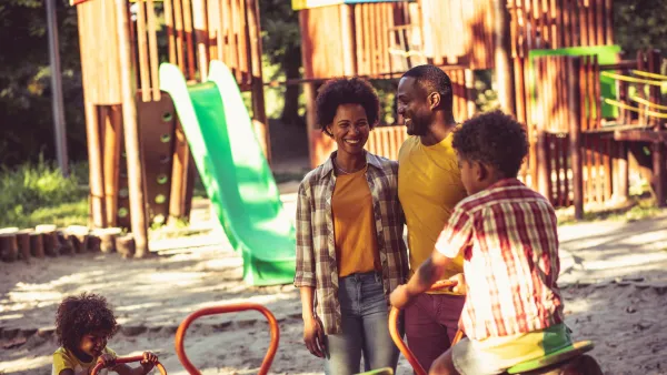 Two African American parents stand on playground with a young boy and toddler girl, looking happy on a sunny day.