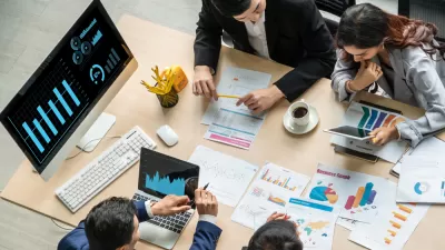 Overhead view of people at a large table working with charts and graphs on laptops and on paper