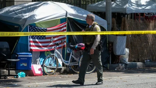 Sheriff's officer walks past tent with American flag draped on front in homeless encampment behind yellow police tape.