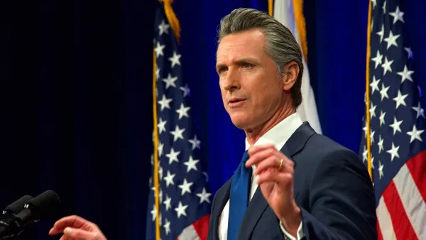 Gov. Gavin Newsom speaks with both hands half-raised. Two American flags, and the flag of California, are behind him.