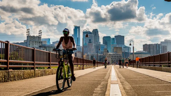 Woman rides bike on bridge in Minneapolis with downtown skyline in background on a sunny day.