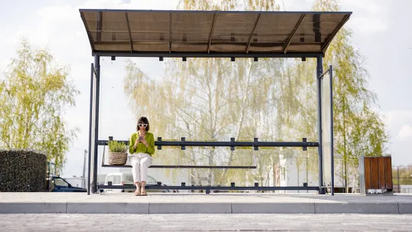 Woman wearing sunglasses sits alone on bench at modern bus stop with shelter and garbage can.