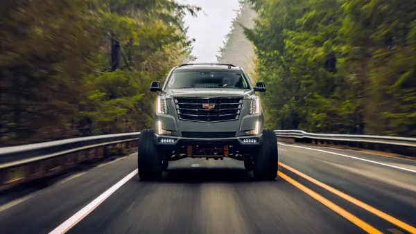 Cadillac Escalade SUV driving toward camera on black asphalt road surrounded by forest