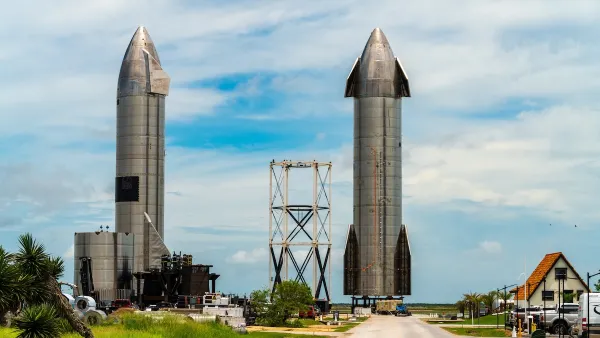 Two large rockets standing upright in Boca Chica, Texas behind Starbase sign with small older home in foreground.