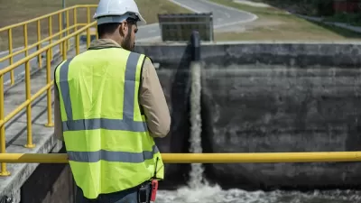 Worker in yellow vest and white hard hat looking down at water basin at wastewater treatment plant