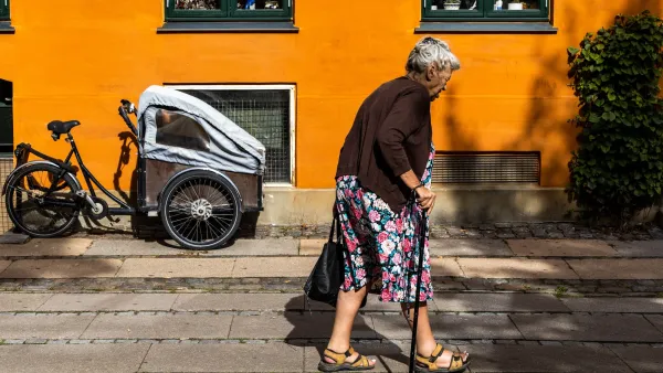 A senior woman with a cane walks by an old house with an ochre and orange color.