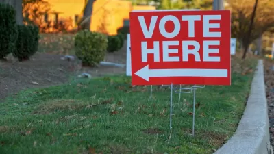 Red "Vote Here" sign with white text and arrow pointing left on green lawn next to curb