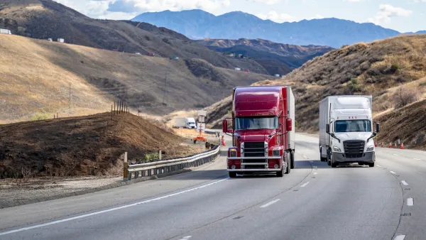 Red and white big rig trucks driving up California highway with rolling dry hills in background.