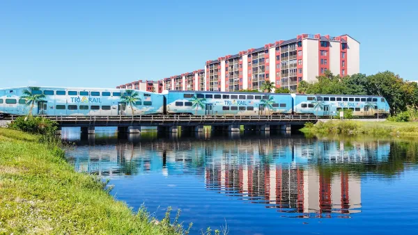 Blue Tri-Rail commuter train passing over small bridge over water with apartment building in background on a sunny day in Florida.