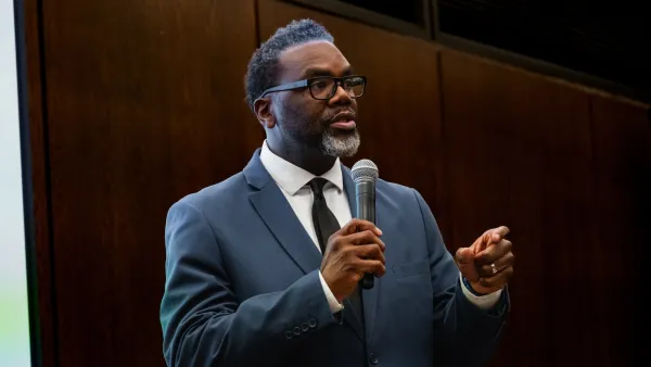 Chicago Mayor Brandon Johnson wearing dark suit speaking into a microphone in front of a dark wood wall.