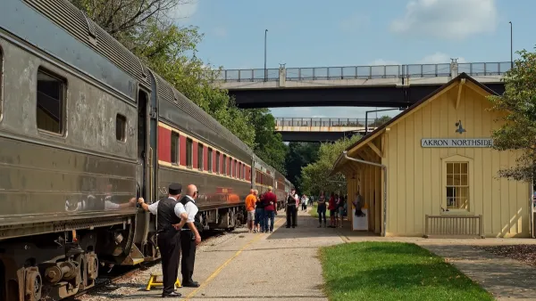 Passengers and crew stand outside of a train. there is a bridge ahead of them and a yellow A frame building.