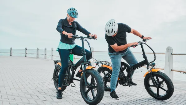 Older couple wearing helmets on electric bikes stopped on a pier next to ocean.