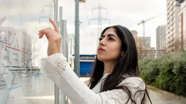 Young woman in white jacket looking at transit map outdoors with hand raised pointing to map.
