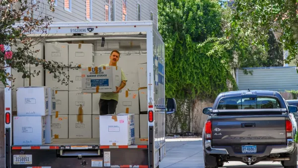 Man standing in moving truck stacking boxes on street with grey truck driving away.