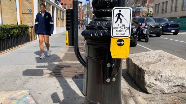 Yellow sign for audible crossing signal on post at intersection in New York City with one person walking on sidewalk in background.