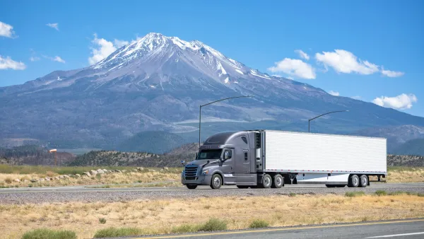 Grey big rig truck with white trailer driving on California highway with snow-capped peak visible in packground against blue sky.