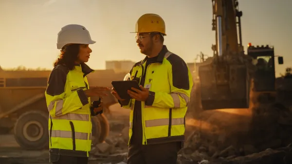 Man and woman in hard hats and safety vests looking at plans on tablet at golden hour at construction site.