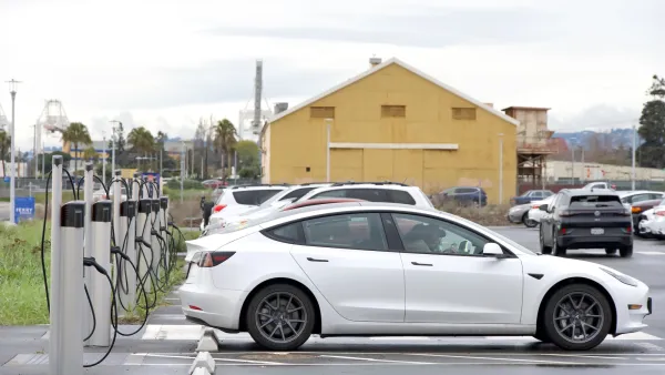 White electric car charging at a station in a parking lot with a yellow barn in background.