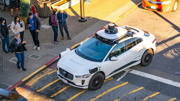 White Waymo self-driving car at intersection in San Francisco with pedestrians taking photos from sidewalk.