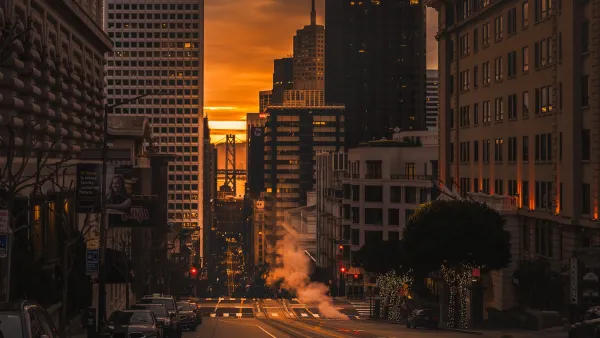 View down a San Francisco street with tall offive buildings on either side and a golden sunrise on the horizon.