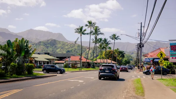 A street with cars and shops, with green mountains in the background. 