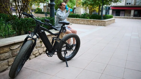 Black electric bike parked on urban walkway with rider sitting on ledge drinking water behind it.