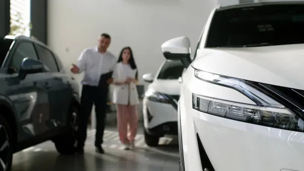 Close-up on white sedan in showroom with salesperson and client standing in background, blurred.