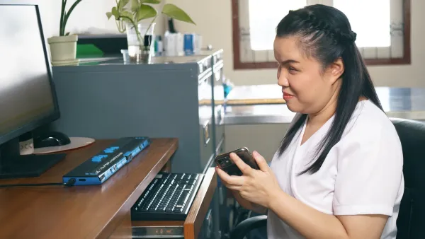 Visually impaired woman in white shirt sits at computer using screen reader device.