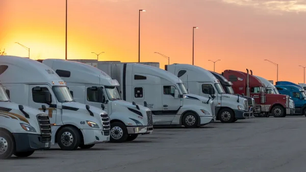 Row of mostly white and one red truck parked in a parking lot at sunset.