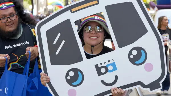 Person in bucket hat and sunglasses holding cardboard cutout of cartoon BART train in parade in San Francisco.