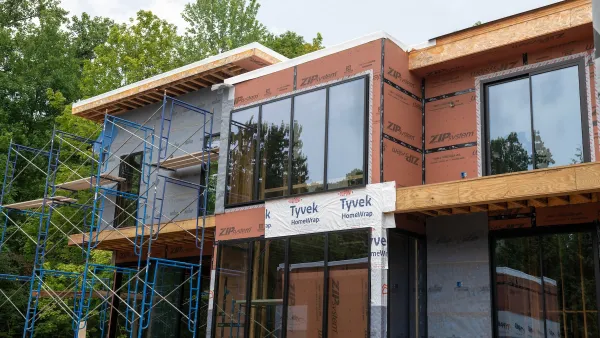 Two-story home or multifamily housing with large windows under construction in front of mature green trees.