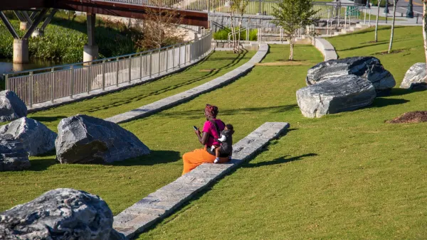An African American woman with a baby on her back sitting in the park surrounded by lush green trees, grass and plants with blue sky at Rodney Cook Sr. Park in Historic Vine City in Atlanta, Georgia.