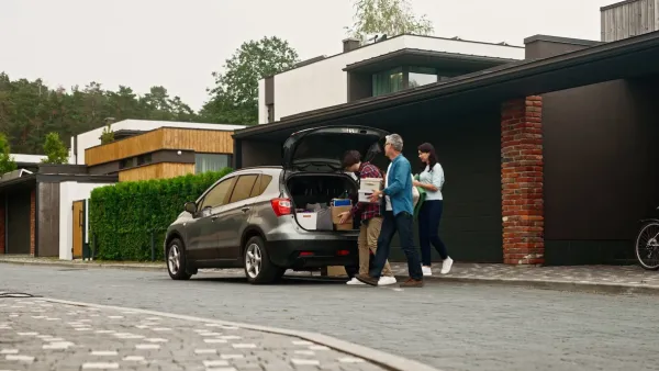 Parents helping child carry boxes to or from car trunk in front of house.