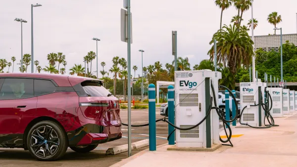 Red electric car plugged into charging station in outdoor parking lot with palm trees in background.