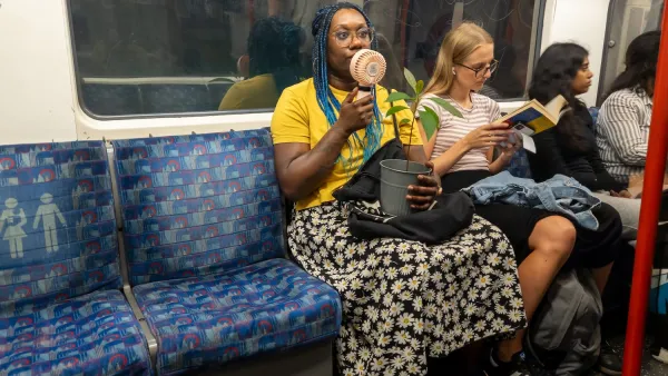 Woman sitting in subway car holds up small portable fan to cool herself.