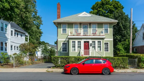 Green two-story wood house in Brighton, Massachusetts on a sunny day with small red sedan parked in front.