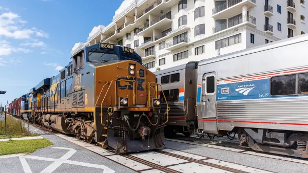Amtrak train next to freight train at a station next to multi-story apartment building.