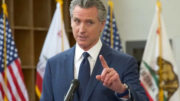Gov. Gavin Newsom speaking behind a podium, pointing a finger upwards. The American and California flags are behind him.