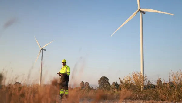 Worker in yellow safety jacket standing in field next to wind turbine.