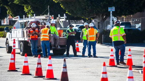 Group of workers in yellow and orange safety vests and hard hats stand in street in Los Angeles next to work truck protected by row of orange cones.