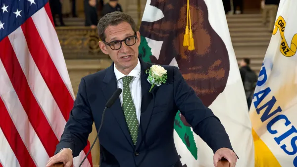 Daniel Lurie, in a suit and green tie, speaks behind a podium with the American flag in the background.