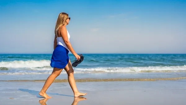Woman in white top walks on beach holding her sandals on sunny day.
