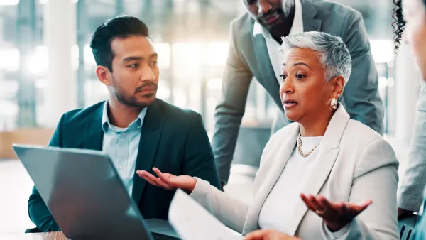 Man and woman in business meeting looking at laptop with another man leaning over desk.
