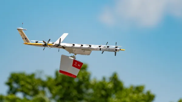 White drone holding red and white delivery box flying over green treetops.