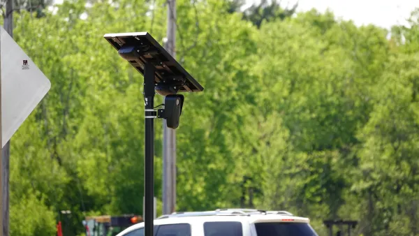 A black Flock traffic camera with solar panel above it installed on a pole outdoors with car visible in background.