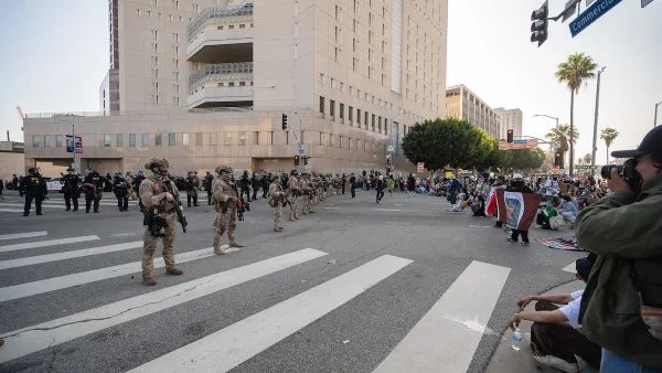 National Guard troops in uniform lined up on a street in downtown Los Angeles next to high-rise jail building with protesters facing them across the street.
