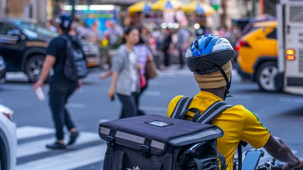A person in NYC bikes behind a bus. They have a helmet on top of a yellow hat and wear a yellow shirt.