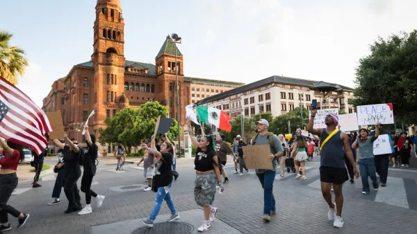 Protesters carrying signs and Mexican and American flags march in San Antonio street against ICE raids with historic Spanish-style building in background.
